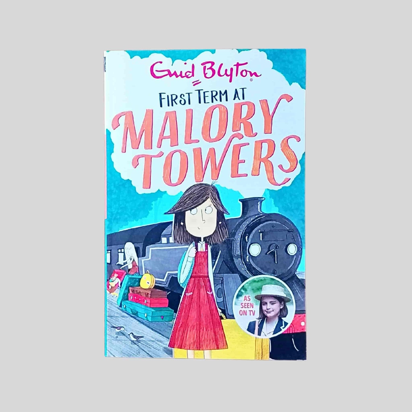 The front cover of the book called First Term at Malory Towers by Enid Blyton showing an illustration of a schoolgirl standing on a railway platform beside a steam train