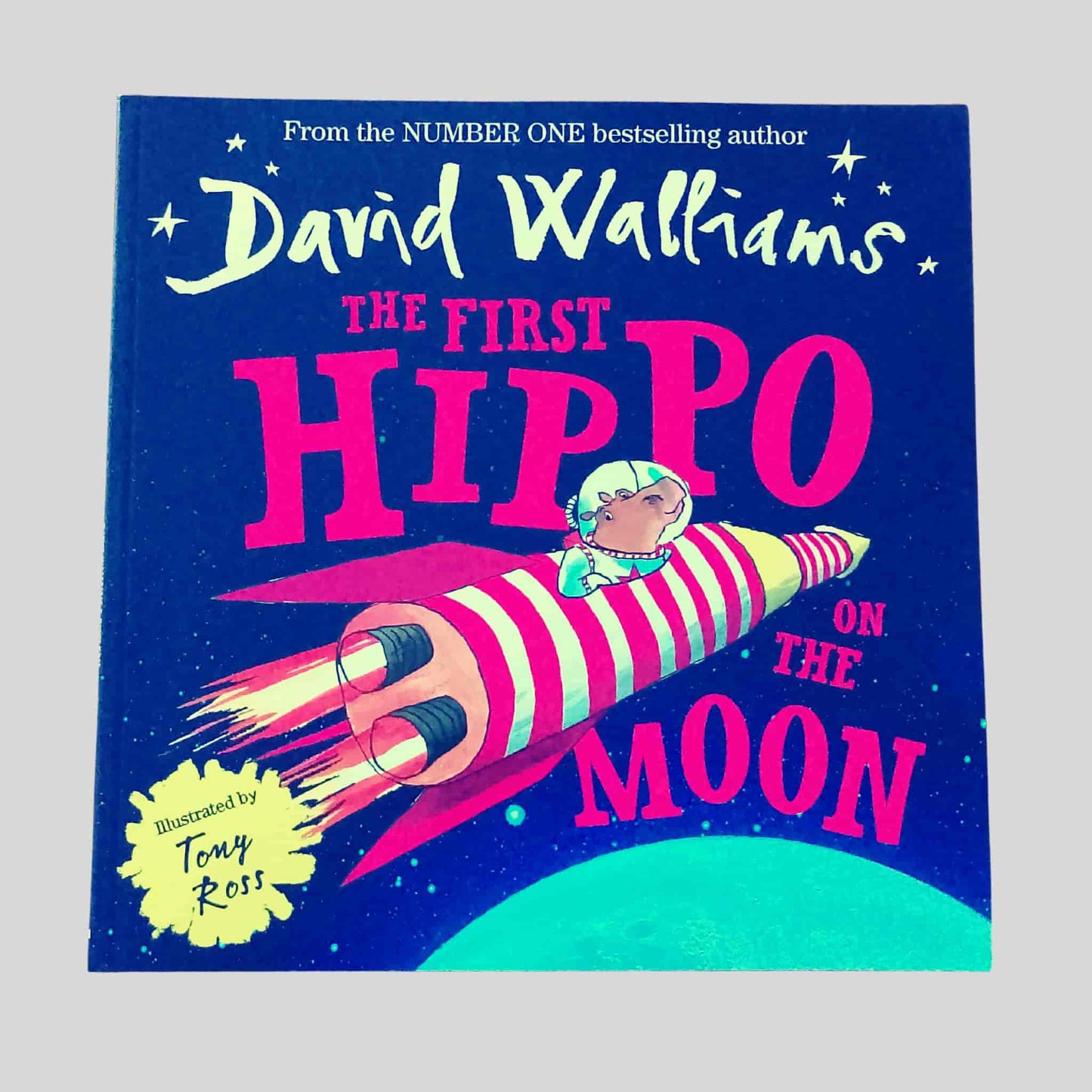 The front cover of the children's picture book The First Hippo on the Moon by David Walliams, illustrated by Tony Ross, showing a hippo astronaut riding a striped rocket through space