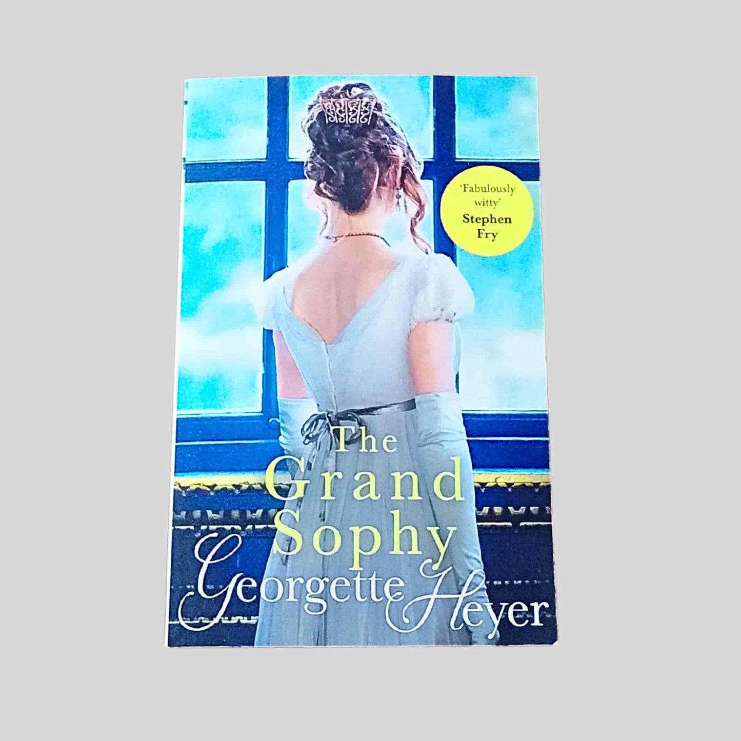 Front cover of The Grand Sophy by Georgette Heyer, paperback edition, showing a Regency-era woman in a pale blue gown standing with her back to the viewer at a large window, evoking wit and high-society romance