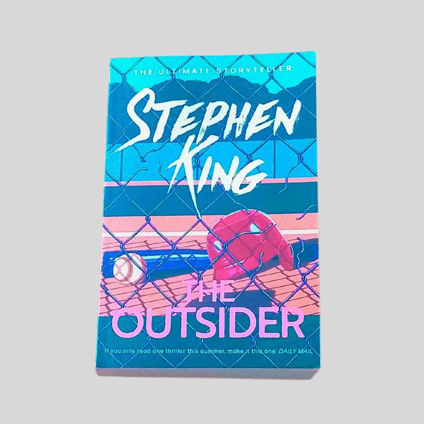 Front book cover of The Outsider by Stephen King, paperback edition, showing a chain-link fence with a baseball and helmet on a pitch behind it