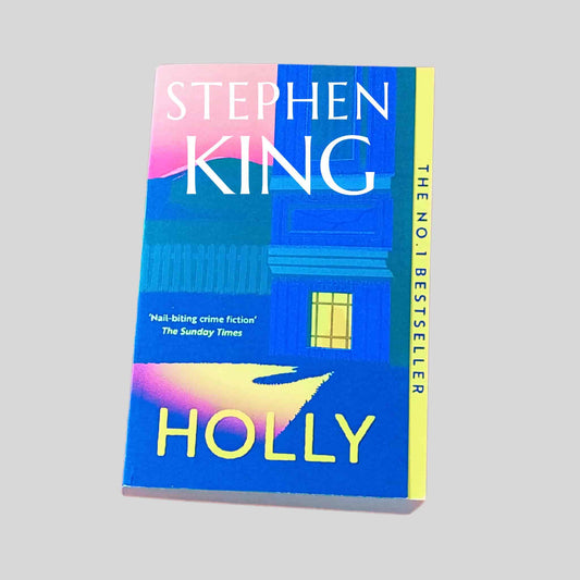 The front book cover of Holly by Stephen King in paperback, showing a blue illustrated house at night with a yellow lit window