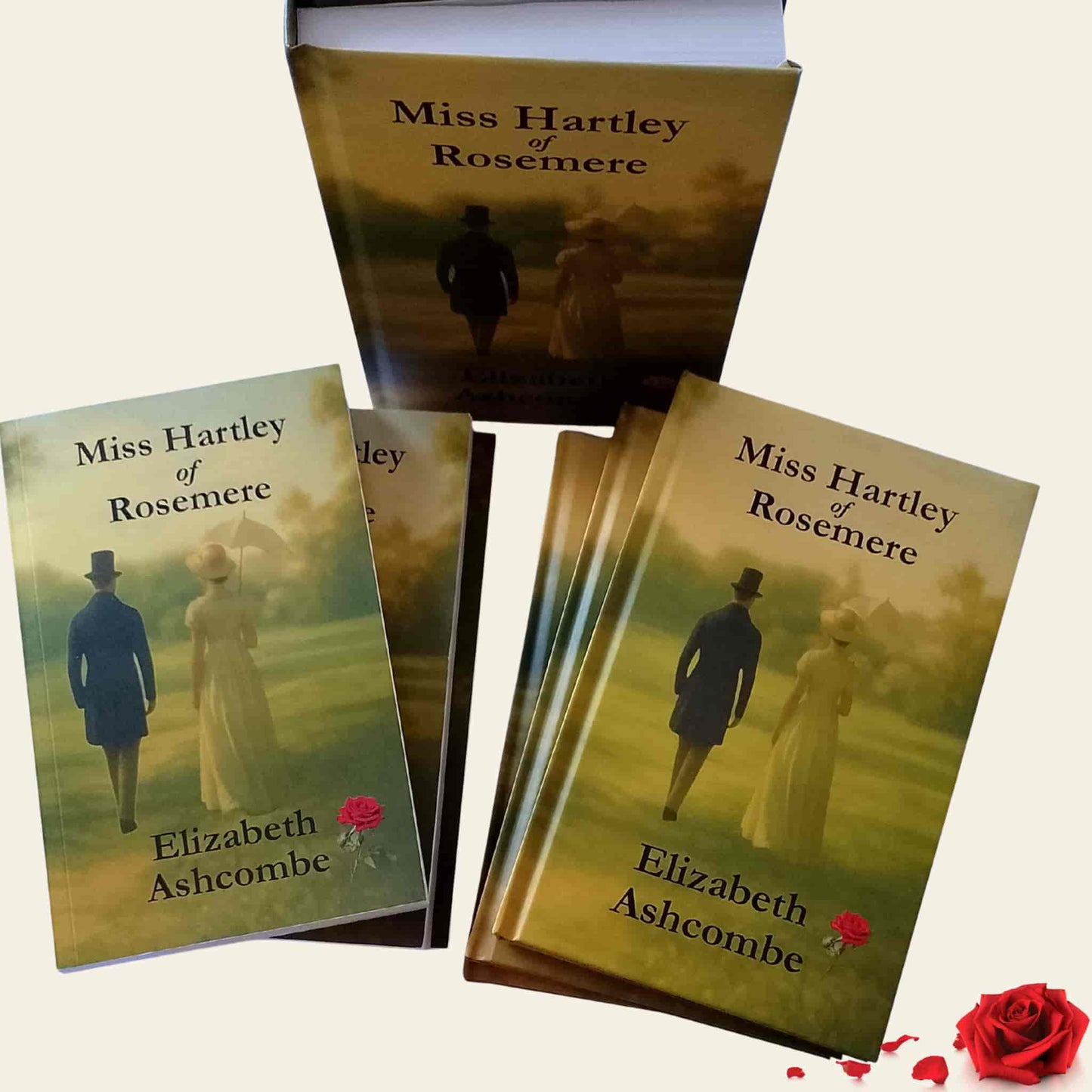 A display of hardback and paperback editions of the book Miss Hartley of Rosemere by Elizabeth Ashcombe, the cover features a Regency-era couple viewed from behind walking together in a countryside setting, with a red rose placed at the bottom right next to the books