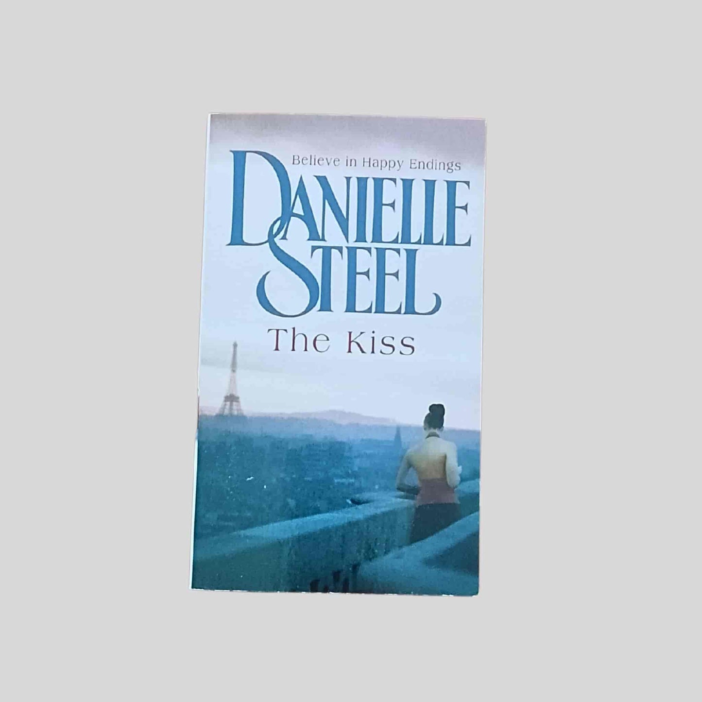 Paperback book cover of The Kiss by Danielle Steel, showing a woman standing on a Paris balcony overlooking the city with the Eiffel Tower in the distance